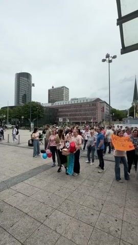 The video shows a crowd of people gathered in an outdoor urban plaza on a cloudy day. The people are of various ages and genders, and many are dressed casually. Some individuals are holding signs and balloons. The signs are mostly handwritten and some appear to be promotional or celebratory in nature. One sign clearly visible reads "WELCOME TO GRIE". Several balloons are visible, predominantly red, white, and blue. In the background, there are several multi-story buildings, one of which appears to be a modern office building, and another has a distinct spire, suggesting a church or similar structure. The ground is paved with large square tiles. People are standing, walking, and interacting within the crowd. The overall atmosphere appears to be one of a public gathering or event.