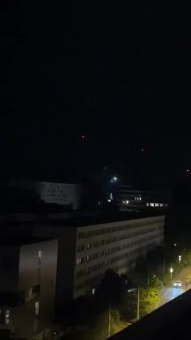 The video shows a nighttime cityscape with multiple buildings. Two red lights are visible in the dark sky, possibly aircraft. A small burst of fireworks briefly illuminates the sky with a white and pink explosion. A few lights are on in the buildings, and streetlights illuminate a road with a moving vehicle.