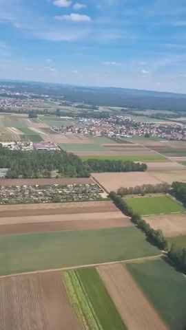 The video shows an aerial view of a landscape with fields, trees, buildings, and a town in the distance. The fields are a mix of green and brown, possibly indicating different crops or stages of growth. There are patches of trees scattered throughout the landscape, and a distinct line of trees bordering some fields. Several buildings, possibly industrial or commercial complexes, are visible near the fields. In the distance, a town with clustered houses can be seen. The sky is blue with scattered white clouds.