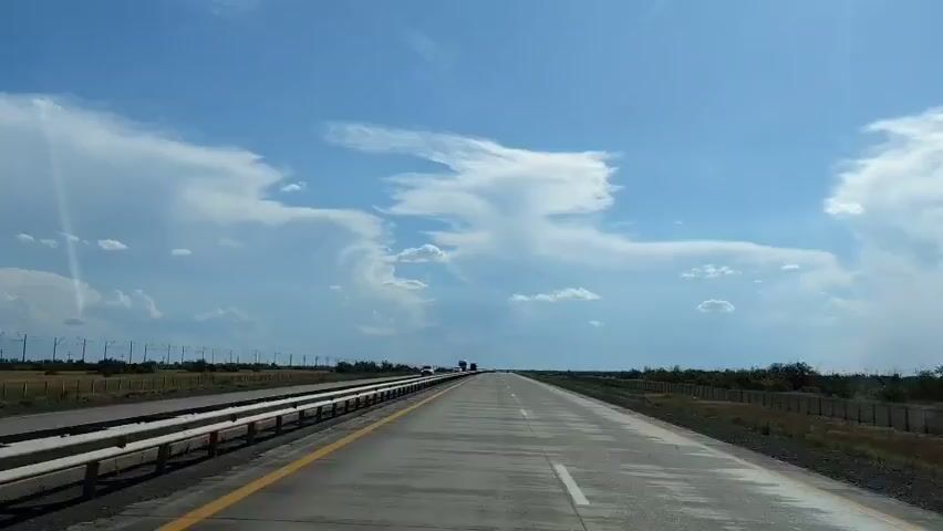 A straight, paved highway stretches into the distance under a bright blue sky with scattered white clouds. On the left side of the highway, a metal guardrail separates the road from a grassy shoulder. Beyond the shoulder, a fence runs parallel to the road, with sparse vegetation and a few trees visible. Several vehicles, including cars and trucks, are traveling on the highway in the same direction as the camera. The overall scene suggests a daytime journey on an open road in a rural or semi-rural environment.