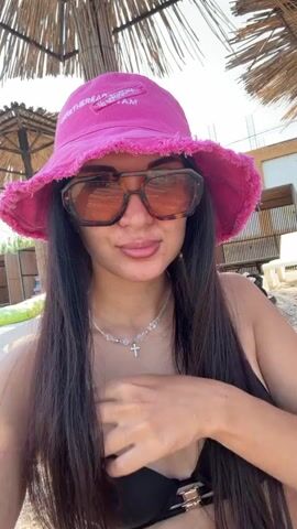 A young woman with long dark hair, wearing a pink bucket hat, sunglasses, a black bikini top, and a necklace with a cross pendant, is taking a selfie on a beach. She is sitting under a thatched umbrella, and in the background, there are more beach umbrellas, lounge chairs, and some buildings. The lighting suggests it is daytime.
