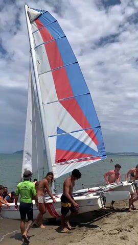 A group of people are on a sandy beach, pushing a small sailboat into the water. The sailboat has a tall mast with a sail that is red, white, and blue. Several people are in bathing suits, and some are wearing life vests. The water is calm, and the sky is cloudy. There are other sailboats in the background.