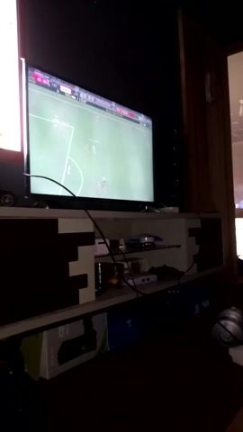 The video captures an indoor scene, likely within a home in Kecamatan Pacet, Indonesia, focusing on a gaming setup. The room is dimly lit, suggesting either evening or a deliberate darkening of the space for an optimal viewing experience. A large flat-screen television is prominently displayed on the left, showing a football (soccer) video game in progress.

To the right of the television, a wooden shelf or cabinet is cluttered with various electronic devices, including what appears to be components of a gaming console, and a tangle of cables. As the video progresses, two young men are seen in the frame. One young man, dressed in a white collared shirt and dark trousers, squats down in front of the gaming console. He holds a game controller, appearing focused and engaged as he interacts with the console, presumably setting up or playing the football game on the screen. The legs of a second person, also squatting, are briefly visible on the right, indicating shared participation or observation of the gaming session.

In the background, an open doorway offers a glimpse into an adjacent room, which seems to be another part of the living space. A white household appliance, possibly a fan or air purifier, is situated on the floor near the gaming area. The overall scene depicts a casual leisure activity centered around video gaming. No specific text or branding is clearly legible on the screen other than the general interface of the football video game.