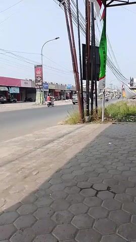 The video captures an outdoor scene on a sunny day. The camera is positioned on a sidewalk paved with hexagonal tiles, looking out at a paved road. On the left side of the road, there is a row of low-rise commercial buildings with signs, some of which are in red and white. A few motorcycles are visible on the road, some with riders. There are telephone poles and wires overhead, casting shadows on the sidewalk. A green and white flag is hanging from one of the poles. The overall environment appears to be a commercial or suburban area.