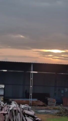 The video shows a time-lapse of a cloudy sky with the sun partially visible, casting a warm glow. The landscape transitions between what appears to be a construction site with a covered area, fencing, and building materials, to a more natural setting with trees and vegetation. There are no people visible in the footage. The camera appears to be moving, possibly from a vehicle.