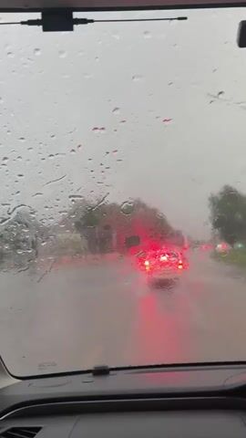 The video shows a view from inside a car looking out the windshield. Rain is falling, creating streaks and droplets on the glass. The road ahead is wet and appears to be flooded in some areas. Several cars are visible in the distance, their taillights illuminated. Trees and foliage can be seen on the sides of the road. The sky is overcast, contributing to the gloomy atmosphere. The interior of the car, including the dashboard and windshield wipers, is partially visible in the foreground. The overall scene depicts driving in heavy rain.
