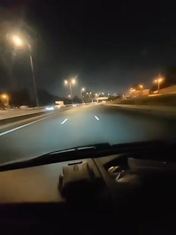The video shows a first-person view from inside a car at night, driving on a highway. The road is well-lit by streetlights and the headlights of the car. Other vehicles are visible in the distance on the highway. The dashboard of the car is visible in the foreground. The overall environment is a typical highway scene at night.
