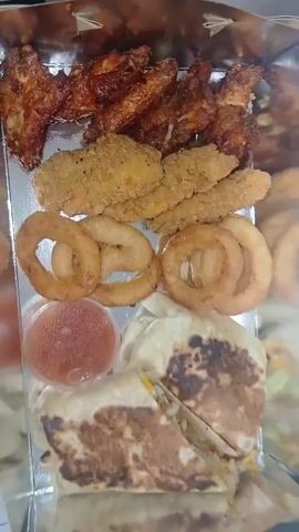 The video is a close-up overhead shot of a platter of fried food. From top to bottom, the platter contains chicken wings, chicken tenders, onion rings, a small plastic container of sauce, and a quesadilla that has been cut in half to reveal the filling. The lighting appears to be artificial and is causing some glare on the food and the platter.