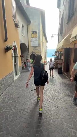 The video captures a bustling street scene in Malcesine, Italy. A young woman with a backpack is walking away from the camera down a cobblestone street lined with buildings. She is wearing a dark grey t-shirt, black shorts, and bright yellow sneakers. In front of her, a woman in a patterned dress and a straw hat walks with another person. To the right, an older woman in a white t-shirt and shorts walks towards the camera.

The buildings on either side of the street have a rustic, aged appearance. One building features a large poster with an image of Gustav Klimt. The poster appears to be advertising an exhibition, with text including "GUSTAV KLIMT" and dates that suggest it's a contemporary event.

The street opens up to a glimpse of a body of water and distant hills, indicating the proximity of a lake or sea. The sun is shining, suggesting a warm, pleasant day, likely midday or early afternoon given the brightness. The overall atmosphere is one of a lively tourist town, with people enjoying a stroll through its charming streets.