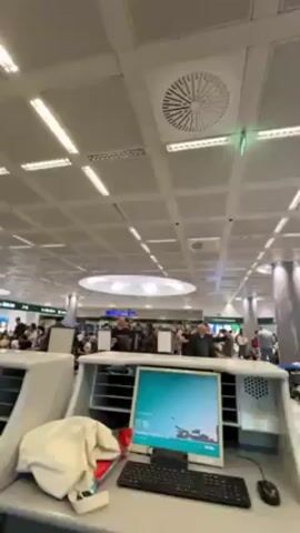 The video shows the interior of a busy airport. The camera pans from a check-in counter with a computer, keyboard, and miscellaneous items on it, across a crowded area with many people waiting in line. Several airport staff in uniform are visible. The camera then moves towards a glass door with a green evacuation plan sign on it. The overall environment is well-lit with a grid-patterned ceiling and fluorescent lighting.