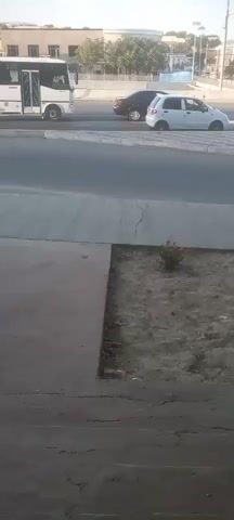 A bus is visible on the left side of the screen, parked at what appears to be a bus stop. A dark-colored sedan is driving in the lane next to the bus, followed by a white hatchback. The cars are moving from left to right across the screen. The background consists of buildings and trees under a clear sky. The foreground shows a concrete sidewalk and a patch of dirt with some sparse vegetation. The scene appears to be a typical urban street environment during daylight hours.