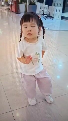 A young child, likely a toddler, stands in what appears to be a public indoor space with tiled flooring. The child has dark hair pulled into two pigtails and is wearing a white t-shirt with a light-colored graphic and light pink, baggy pants. The child is facing forward, with their left leg slightly bent and their right leg extended. Their left arm is raised with the palm facing outwards, as if waving or holding up a hand. The child's eyes are closed, and their mouth is slightly open, giving the impression they are singing or speaking. The background shows a polished floor with reflections, a large potted plant, and some blurred figures and objects in the distance, including what looks like shopping carts. The lighting is bright and even.
