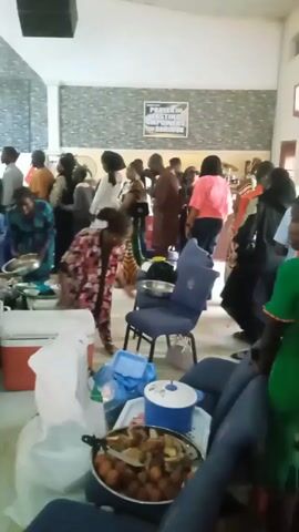 The video shows a gathering of people in a large indoor space, possibly a church hall or community center. Several individuals are seen getting food from serving tables, indicating a meal or reception is taking place. There are platters of food, containers, and bags near chairs in the foreground, suggesting that people are serving themselves or preparing to eat. The crowd is diverse, with people wearing various styles of clothing. Some individuals are moving around, while others are standing in line for food. The environment seems lively, with conversations and movement throughout the space.