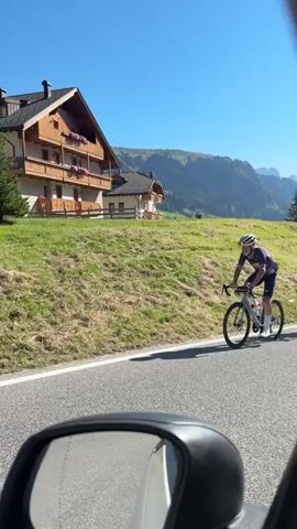 The video begins with a wide shot of a rural road winding through a green, hilly landscape under a clear blue sky. Two cyclists, wearing racing attire, are seen riding on the road. The first cyclist, in the foreground, is wearing a blue and red patterned jersey and black shorts, riding a grey bicycle. The second cyclist, further up the road, is wearing a black jersey and shorts, riding a green bicycle. In the background, traditional European-style houses with wooden balconies and flower boxes are nestled on the hillside, with a backdrop of majestic mountains. The camera perspective appears to be from a vehicle traveling behind the cyclists, as a car's rearview mirror is visible in the bottom corner of the frame. The overall scene depicts a picturesque cycling journey through a serene, mountainous region.