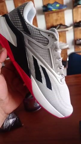 A close-up video showcases a hand holding and rotating a white and light grey sneaker with black accents and a vibrant pink/red sole. The shoe appears to be a Reebok Nano X2, with "Reebok" and "NANO X2" faintly visible on the side. The hand turns the shoe to display its front, sides, and heel, highlighting its design and sole pattern. In the background, numerous other athletic shoes are neatly displayed on wooden shelves, suggesting a retail environment such as a shoe store. The shoe is being held over a wooden table, next to a small can and a black plastic bag. The scene takes place in Bireuenmeunasahcapa, Indonesia.