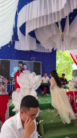 This video captures a lively outdoor celebration, likely a wedding or engagement party, taking place under a large tent adorned with white and blue fabric drapes and string lights.

In the foreground, a man in a white shirt smiles and laughs, appearing to enjoy the event. On a small raised stage with green carpet, a woman in a light-colored, flowing dress (possibly the bride) dances with visible joy and enthusiasm. Two large white paper lotus flowers are positioned on the stage near her.

Behind the dancer, a woman in a red dress sings into a microphone, leading the musical entertainment. To her right, a band plays, with a man on a keyboard and another on a guitar. A large screen in the background displays a photo of a couple, reinforcing the celebratory theme.

On the left side of the frame, a table covered with a red tablecloth holds a tiered cake and a decorative birdcage-like object, along with floral arrangements. The overall atmosphere appears festive and happy, characteristic of a wedding celebration in Tay Son Commune, Vietnam.