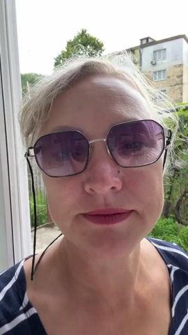 A woman with blonde hair and sunglasses smiles at the camera. She is wearing a navy and white striped shirt. The background shows greenery and a building. It appears to be daytime and the weather is clear. The woman is outdoors, possibly on a porch or patio, given the white railing visible on the left side of the frame. Her expression is cheerful and she has dimples.