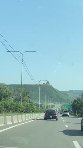 A vertical, POV video captures a view from inside a vehicle traveling on a highway. The highway has multiple lanes separated by a concrete barrier and a white line. Streetlights line the right side of the road. On the left side, a lush green hill rises, topped by an ancient stone structure that appears to be a monastery or church with a domed roof. The sky is clear and blue. Several cars are visible on the highway, including a black hatchback directly ahead and a white SUV further down the road. A green highway sign with white lettering and a parking symbol is visible on the right. Trees and other vegetation border the highway. The video appears to be filmed during daylight.