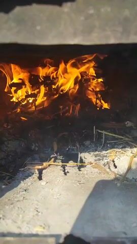 The video shows a fire burning inside a stone structure. The flames are bright orange and yellow, consuming small pieces of wood and other materials. The fire is contained within a gray stone box, and the bottom of the structure is covered in ash and unburnt debris. The camera angle is looking directly into the fire from the outside.