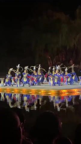 The video shows a group of performers on a stage above water at night. The performers are dressed in blue and gold costumes and have feathered headdresses. They are drumming on small drums. The stage is lit with yellow lights and the reflections can be seen on the water. The video is taken from behind some audience members. There are trees and foliage in the background.