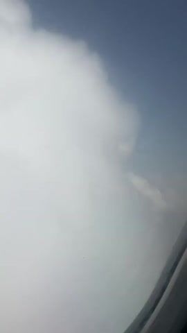 The video shows a view from an airplane window. The sky is partially clear with fluffy white clouds. As the video progresses, the plane descends, and the landscape below becomes visible. It appears to be a residential area with buildings and greenery. The airplane window frame is visible in the bottom right corner of the frame.