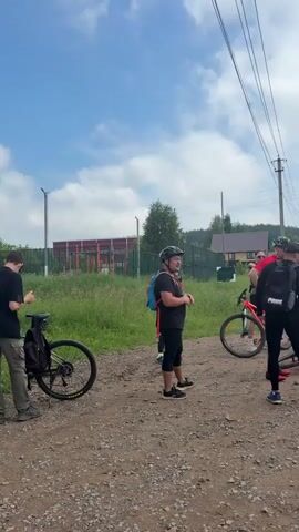 A group of people wearing athletic clothing and helmets are gathered around bicycles on a dirt path. The sky is partly cloudy. In the background, there is a fence, grass, trees, and buildings. Some people appear to be talking to each other, while others are looking at their bicycles. One person is adjusting their helmet. Another person is holding a bicycle. A car is parked nearby.
