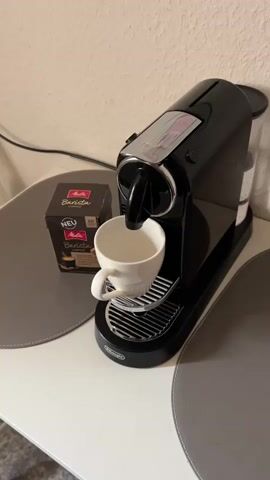 The video shows a close-up, slightly high-angle shot of a black DeLonghi Nespresso coffee machine. A white ceramic cup is positioned on the drip tray, directly beneath the coffee spout. Coffee is actively brewing and flowing from the spout into the cup, creating a light brown stream. To the left of the coffee machine, there is a small, dark-colored cardboard box of "Barista" coffee capsules, with a "NEU" (New) label visible. The coffee machine and cup are placed on a white surface, likely a countertop or table, with two large, oval-shaped grey placemats partially visible on either side. The background is a plain, light-colored wall. A green indicator light is illuminated on the top-front of the coffee machine.