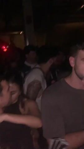 The video depicts a crowded indoor space, likely a club or party, with many people moving and dancing. The lighting is dim and shifts in color, suggesting a dynamic atmosphere. Individuals are seen wearing various outfits, and some appear to be conversing or interacting closely with one another. The environment includes architectural features suggesting an upper level or balcony. The camera movement is unsteady, giving a sense of immersion within the crowd.