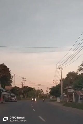 The video shows a road scene. The camera is positioned on a moving vehicle traveling on a paved road with white lane markings. Several cars and a motorcycle are visible, moving in the opposite direction. Utility poles line the sides of the road, with many power lines crisscrossing above. Vegetation, including trees and bushes, is visible on either side of the road. Buildings are scattered alongside the road, with one structure appearing to be a small shop or vendor stand. The sky is a mix of light blue and pale orange hues, suggesting early morning or late afternoon light. The bottom of the video displays the text "OPPO Reno8" and the date "2026.05.23 06:34".