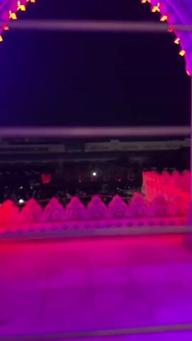 Here's a description of the video:

The video shows a nighttime view of a fountain display with people gathered around it, taken from a high vantage point. The foreground shows an ornate, pink-lit balcony ledge with a decorative pattern. Beyond the ledge, there's a crowd of people watching the fountains. The fountains shoot water in vertical streams, lit up with changing colors. Buildings are visible in the background, illuminated with lights. A bright light sweeps across the scene horizontally. Music plays in the background.