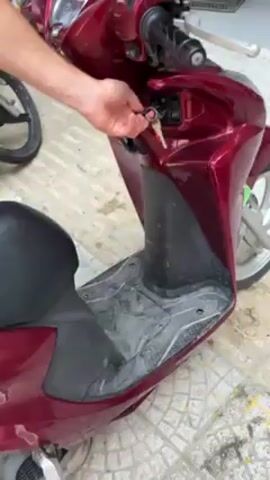 A person is inserting a key into the ignition of a red scooter. The person turns the key, presumably starting the scooter. The person then places a white and orange helmet on the seat of the scooter. The scooter is parked on a tiled surface outdoors. The person is wearing shorts. A bicycle is visible in the background.