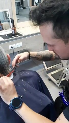 In a medium shot taken from behind, a person with silver hair receives a haircut from a hairdresser in a salon. The hairdresser, who is predominantly visible, has dark hair with a noticeable fade and a close-cropped beard. He is also sporting a tattoo that covers his arm and is wearing a blue smartwatch on his left wrist. He is diligently cutting the client's hair with a pair of scissors while using a red comb to guide the strands. The client is wearing a black barber's cape and a paper collar underneath. The countertop behind them is decorated with a mirror, a spray bottle, and a jar. Cut hair is scattered over the client's cape.