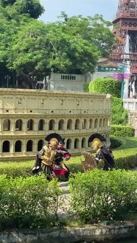 The video shows three people dressed in Roman-style armor and costumes. They are standing in front of a miniature replica of the Colosseum, which is yellow and has arched openings. The ground is covered in green grass and bushes. One person on the left is wearing gold armor, a black cape, and a helmet with a crest. They are holding a sword and a shield. Another person in the middle is wearing a darker, more ornate armor and a helmet, and is also holding a sword, appearing to be in a fighting stance. The third person on the right is wearing a black cape and a helmet with a crest, and is holding a large, round shield with a gold design. They are all positioned as if in a mock combat scenario. In the background, there are trees, a building, and a distant structure resembling the Eiffel Tower, suggesting this is an outdoor miniature park or attraction. The lighting indicates it is daytime, likely sunny.