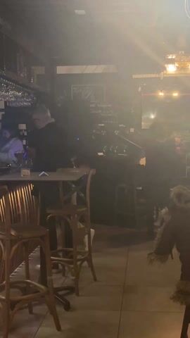 A medium shot video shows the inside of a dimly lit bar with patrons seated at the bar. In the background, the bar is fully stocked with glasses and alcohol, and a beer tap system is also visible. In the foreground, two wooden bar stools sit empty in front of the bar. Two men are standing at the bar, one wearing red sunglasses. The man in sunglasses adjusts his hair. There are several drinks lined up on the bar, including a pink cocktail and several beers. A bartender wearing a hat can be seen near the beer taps. In the background, another patron is seated at the bar. Latin music is audible in the audio.