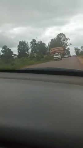 The video shows a view from inside a vehicle traveling on a road. In the distance, a white truck is visible, heavily loaded with what appear to be large, cylindrical bales. Several other vehicles are also on the road ahead of the truck, including SUVs and cars. The environment appears to be rural, with trees and vegetation on either side of the road. The sky is overcast, suggesting a cloudy day. The camera is positioned looking forward, capturing the road and the traffic ahead.