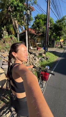 A woman with a long braid, wearing a black sports bra and black shorts, stands beside a bicycle with a red basket on the front. She is holding the handlebars of the bicycle and looking to her left, away from the camera. The bicycle is on a paved road beside a grassy area. Behind her are houses and trees, typical of a residential neighborhood. The video then shifts upward to capture the sun and some buildings. The sky is clear blue.