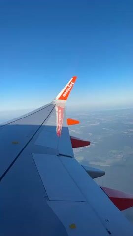 This is a POV video from an airplane window, looking out at the wing. The wing is grey with orange accents and the EasyJet logo visible on the wingtip. The plane is flying over a landscape of green fields and trees under a clear blue sky. The sun is casting a shadow on the wing.