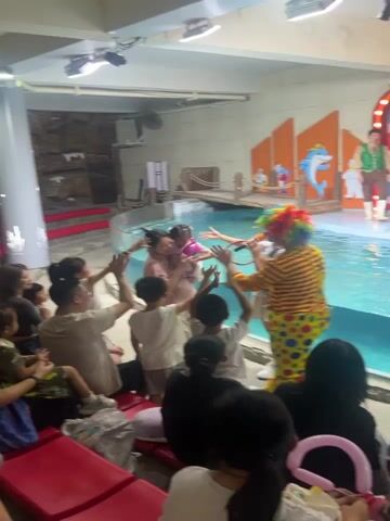 A seal performance is underway in a pool area with an audience watching from bleachers. A young female host, dressed in a vest and skirt, stands by the edge of the pool holding a microphone and a hoop. A trainer in a vest and pants stands on a platform in the pool with a seal. Behind them is a stage decorated with a backdrop featuring a sunburst pattern and Chinese characters that read "Super Performance." A clown in a striped shirt and colorful wig is also present, interacting with the audience. Several audience members, including children and adults, are visible in the foreground, with some raising their hands. The environment appears to be an indoor performance space with artificial rock features visible in the background. The overall atmosphere suggests a lively and engaging show for a family audience.