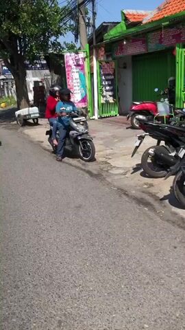 Two people are riding a scooter down a paved road. The person in front is wearing a blue shirt and dark pants. The person behind is wearing a red shirt and dark pants. They are both wearing helmets. The scooter is grey and black. There are buildings on the right side of the road, one of which is painted green. There are several other scooters parked on the right side of the road. There is a trailer attached to a car in the background on the left. The weather appears to be sunny.