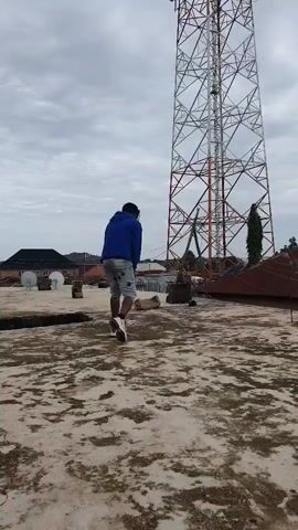 A person wearing a blue hoodie, light-colored shorts, and goggles walks across a rooftop. The person starts by facing away from the camera, then turns to face it. A large radio tower is visible behind the person. The sky is overcast.