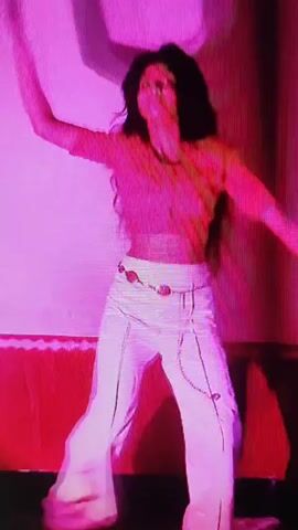 A person with long, dark hair is dancing and singing in front of a pink backdrop. They are wearing a light-colored, possibly crop top and loose-fitting white pants with a decorative belt. The lighting is predominantly pink and purple, with some red accents. The person's movements are energetic and expressive.