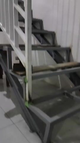 The video shows a metal staircase with white railings. The stairs are made of metal frames with wooden planks as treads. The staircase is located indoors, next to a white wall. There is a brown chair visible under the staircase. The lighting is dim, creating shadows on the stairs and walls. The camera is moving slowly upwards, showing the staircase from a low angle.