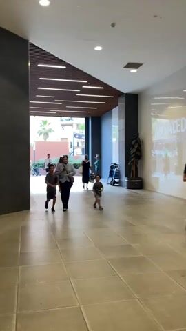 This video captures a moment inside a modern indoor walkway or atrium. The flooring consists of large, square, light-colored tiles. Above, a distinctive architectural feature is visible: a slanted ceiling with a dark wooden finish, segmented by parallel lines of recessed lighting.

In the foreground, two young boys are walking towards the viewer. The boy on the left, wearing a dark t-shirt and shorts, is slightly ahead. The boy on the right, in a lighter t-shirt and shorts, is walking a few paces behind. Between them, a woman wearing a light-colored top and dark pants is walking towards the viewer, looking slightly to her left.

Further back, through an open archway, a group of people are visible. One person, wearing a dark dress, is walking away from the camera. To their right, a person with a stroller is moving.

The environment features a large, dark grey wall on the left, contrasting with a bright white wall on the right, which has a faint reflection or signage visible. A tall, dark potted plant stands near the white wall. The background opens to an outdoor area with buildings and palm trees, suggesting a transition from an indoor to an outdoor space. The overall impression is one of a clean, contemporary public space.