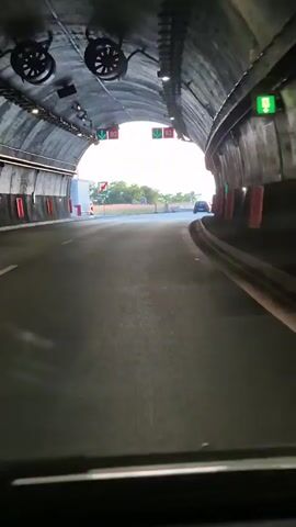 A first-person perspective video shows a vehicle exiting a tunnel and merging onto an open road. The tunnel is illuminated by overhead lights and has signage indicating speed limits. As the vehicle emerges, it is met with a view of a highway with other cars. The sky is cloudy, and there are hills in the distance. The road appears to be asphalt, with clear lane markings. There are guardrails and some vegetation visible along the side of the road. The video captures a brief moment of transition from an enclosed, artificial environment to an open, natural landscape.