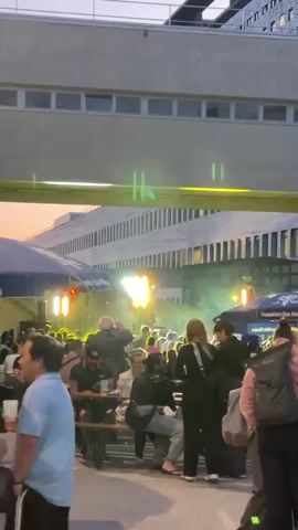 The video shows an outdoor gathering with many people. There is a stage or performance area in the background with bright lights and some smoke or fog. Many people are standing and milling about, and some are seated at tables. There are also umbrellas providing shade, suggesting it might be a market or festival. A building with a distinctive facade and many windows is visible behind the stage area. The lighting suggests it is either early evening or late afternoon. Some individuals in the foreground are blurry due to motion.