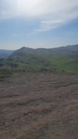 The video presents a scenic, wide-angle view of a natural landscape in Aya, Russia, under a clear, bright sky with scattered white clouds. The camera slowly pans from left to right, revealing an expansive vista. In the immediate foreground is a relatively flat expanse of dark brown, reddish-brown, and gray earth or dirt, possibly a cleared area or a dirt path, with very sparse green growth.

Beyond this foreground, the terrain drops slightly into a valley or lower area, and then rises into a series of rolling green hills and mountains. These hills are covered in lush green vegetation, including grass and scattered patches of trees, primarily deciduous but with some evergreen clusters visible. As the camera pans, more distant mountains come into view, appearing hazy and lighter in color against the horizon. The overall impression is one of a vast and tranquil natural environment, likely captured on a sunny day. No people or visible text are present in the video.