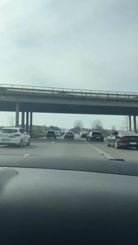 The video shows a view from the dashboard of a car driving on a highway. There are several cars ahead, moving slowly. A concrete overpass is above the car and the other vehicles. The weather appears overcast.