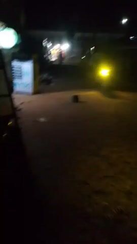 The video shows a nighttime scene on a street. It is dark, with the main light source being streetlights in the distance.  A sign with "NYSC" visible is on the left. Debris is scattered on the ground near a pile of trash to the left of the road.  There is an indistinct object on the road in front of the camera, casting a shadow. Figures and vehicles pass in the distance under the streetlights.