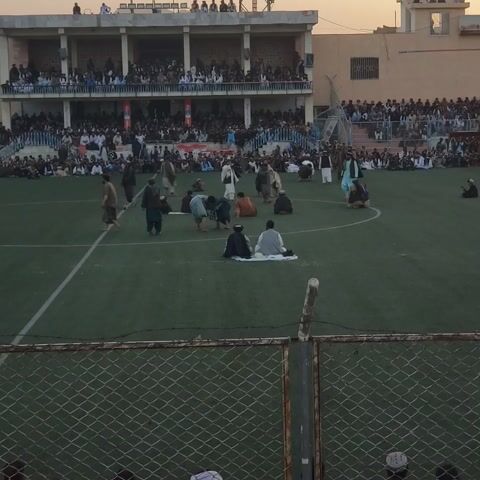 The image shows an aerial view of a soccer field in Kandahar, Afghanistan. The field is artificial turf, and a crowd of people is gathered in the stands on the left side of the frame. Some individuals are also present on the field, many of whom appear to be kneeling or prostrating, suggesting a prayer service or religious gathering is taking place. The time of day appears to be late afternoon or early evening, as indicated by the warm, soft lighting. The weather seems clear. The scene is framed by a chain-link fence on the right. The overall atmosphere seems orderly and communal. There are some visible signs in the background within the stadium structure, but the text is not legible.