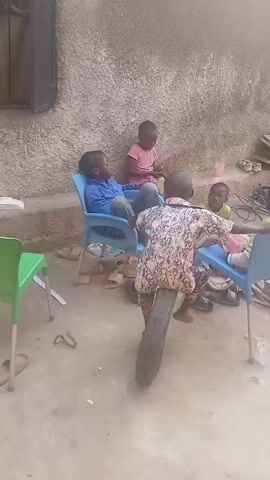 Five young children are in an outdoor area with a gray, textured wall behind them. One child stands facing away from the camera, with a large tire positioned between their legs, as if they are riding it like a motorcycle. Four children are seated in blue plastic chairs around this child. The child in the tire is wearing a patterned shirt and pants. Two of the seated children wear blue and pink shirts respectively, and another child is wearing a green shirt. The last seated child is on the right side of the frame. Other objects are strewn around the area, including sandals, a small green stool, cardboard, and a few wires.